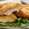 Primo piano di tre medaglioni di patate panati e dorati, disposti su un letto di rucola fresca. Un medaglione è tagliato a metà, rivelando un ripieno generoso di prosciutto cotto e formaggio fuso che fila invitante. La panatura è croccante e granulosa, servita su un piatto di ceramica chiara in un'ambientazione luminosa e moderna.