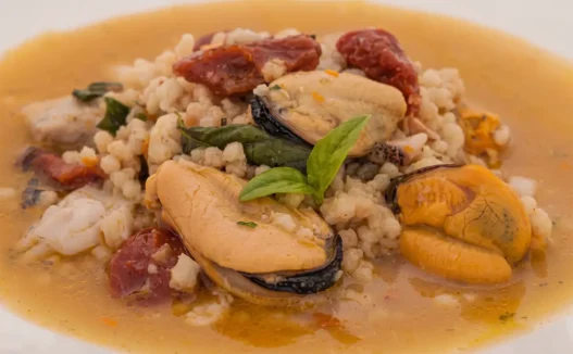 Un piatto di pasta fregola con cozze e pomodori secchi, guarnito con basilico fresco.