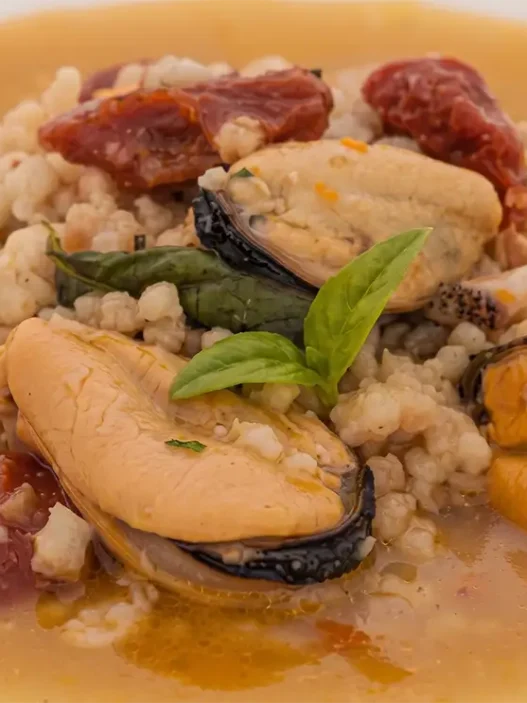 Un piatto di pasta fregola con cozze e pomodori secchi, guarnito con basilico fresco.