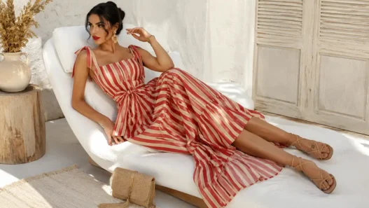 Donna elegante sdraiata su una chaise longue bianca con abito lungo a righe rosse e crema in un ambiente estivo mediterraneo.