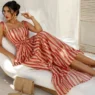 Donna elegante sdraiata su una chaise longue bianca con abito lungo a righe rosse e crema in un ambiente estivo mediterraneo.