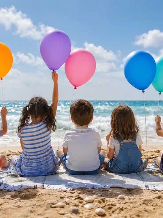 Cinque bambini seduti di spalle sulla spiaggia guardano il mare e tengono in mano palloncini colorati in una giornata di sole.