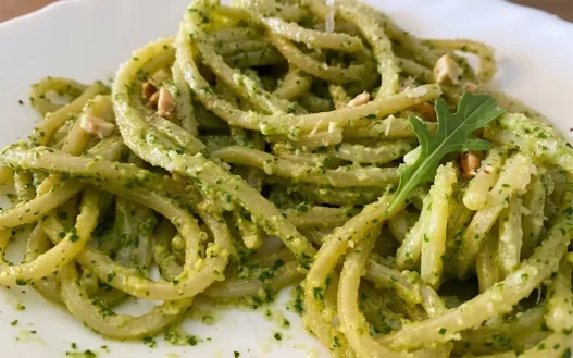 Primo piatto di spaghetti conditi con pesto di rucola e anacardi cremoso, guarnito con granella di frutta secca, scaglie di parmigiano e una foglia di rucola fresca su un piatto bianco.