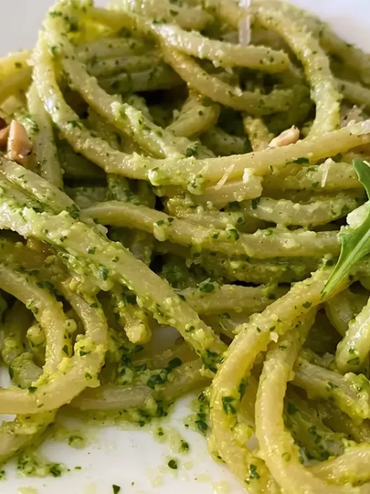 Primo piatto di spaghetti conditi con pesto di rucola e anacardi cremoso, guarnito con granella di frutta secca, scaglie di parmigiano e una foglia di rucola fresca su un piatto bianco.