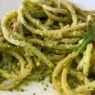Primo piatto di spaghetti conditi con pesto di rucola e anacardi cremoso, guarnito con granella di frutta secca, scaglie di parmigiano e una foglia di rucola fresca su un piatto bianco.