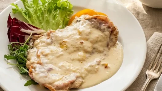 Cotoletta alla Bolognese dorata servita su piatto bianco con prosciutto crudo, formaggio fuso e guarnizione di pomodoro e basilico.