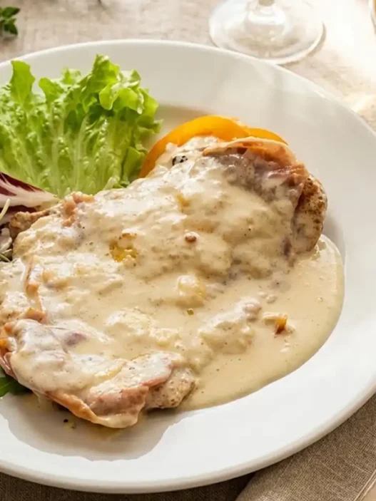 Cotoletta alla Bolognese dorata servita su piatto bianco con prosciutto crudo, formaggio fuso e guarnizione di pomodoro e basilico.