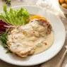 Cotoletta alla Bolognese dorata servita su piatto bianco con prosciutto crudo, formaggio fuso e guarnizione di pomodoro e basilico.