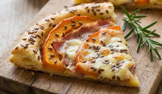 Trancio di sfogliata salata con zucca arancione, fette di speck e taleggio fuso in superficie, cosparso di semi di lino
