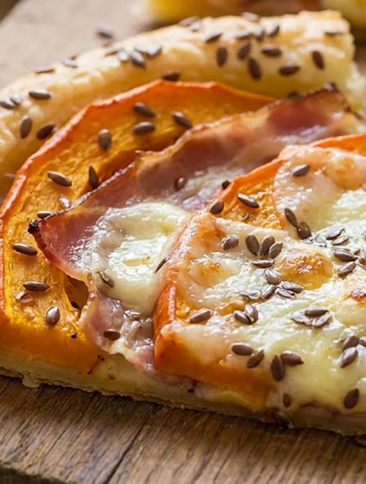 Trancio di sfogliata salata con zucca arancione, fette di speck e taleggio fuso in superficie, cosparso di semi di lino