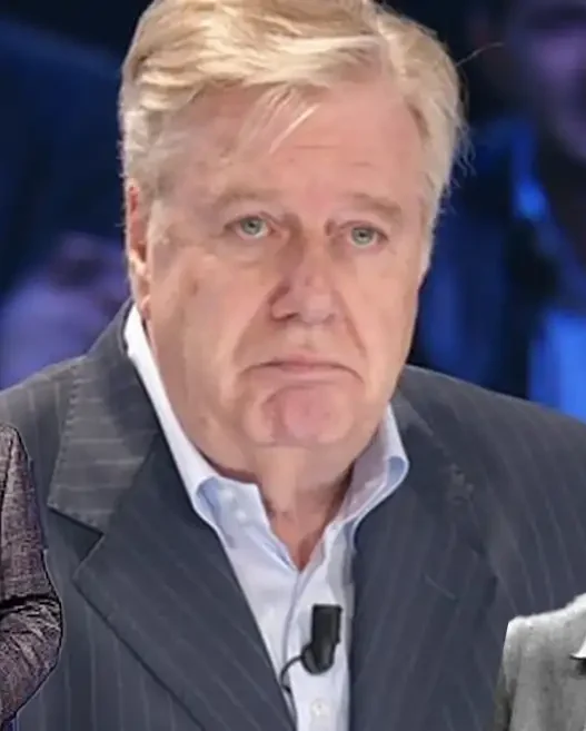 Primo piano di Claudio Lippi sorridente durante una trasmissione televisiva, a contrasto con la notizia del ricovero
