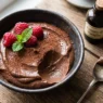 Primo piano di una mousse al cioccolato cremosa e scura con avocado, servita in una ciotola rustica con lamponi e menta.