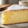 Fetta di Torta Paradiso altissima e super soffice, ricoperta di zucchero a velo, servita su un piatto in ceramica rustica con una tazza di caffè sullo sfondo.