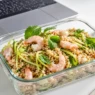 Schiscetta estiva in vetro con insalata di farro, gamberetti e zucchine crude tagliate a julienne, ideale per il pranzo in ufficio