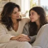 Madre affettuosa tiene la mano della figlia adolescente sul divano, simbolo di supporto psicologico e prevenzione dei disturbi alimentari.