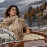 Donna sofisticata in trench e cachemire attraversa il Lago di Como in autunno a bordo di un motoscafo in legno, con ville storiche sullo sfondo.